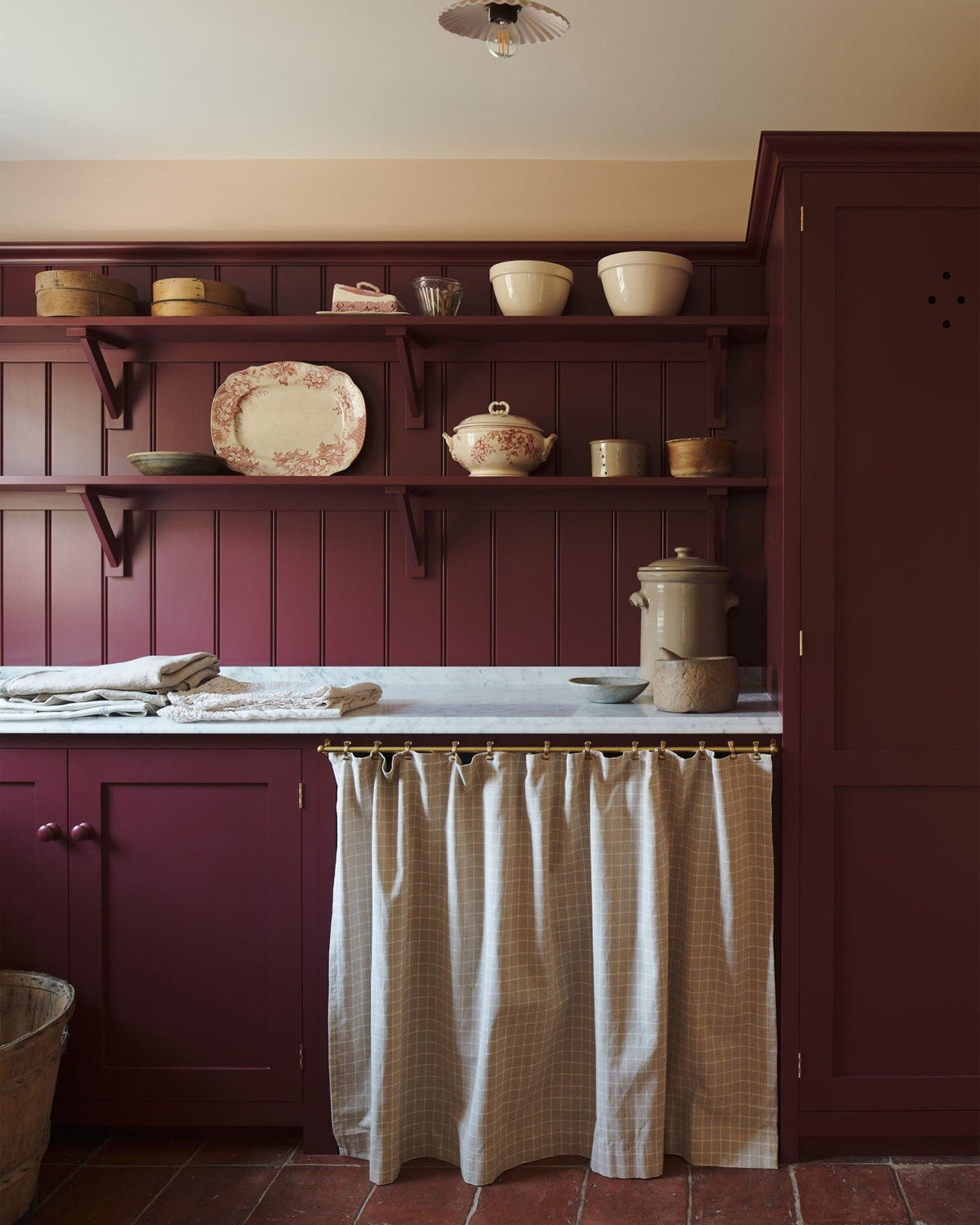 A straight-on view of a red painted Shaker-style utility kitchen with a tall pantry cupboard, open storage behind a red striped curtain and tongue and groove wall panelling and shallow shelves decorated with vintage crockery. TEST
