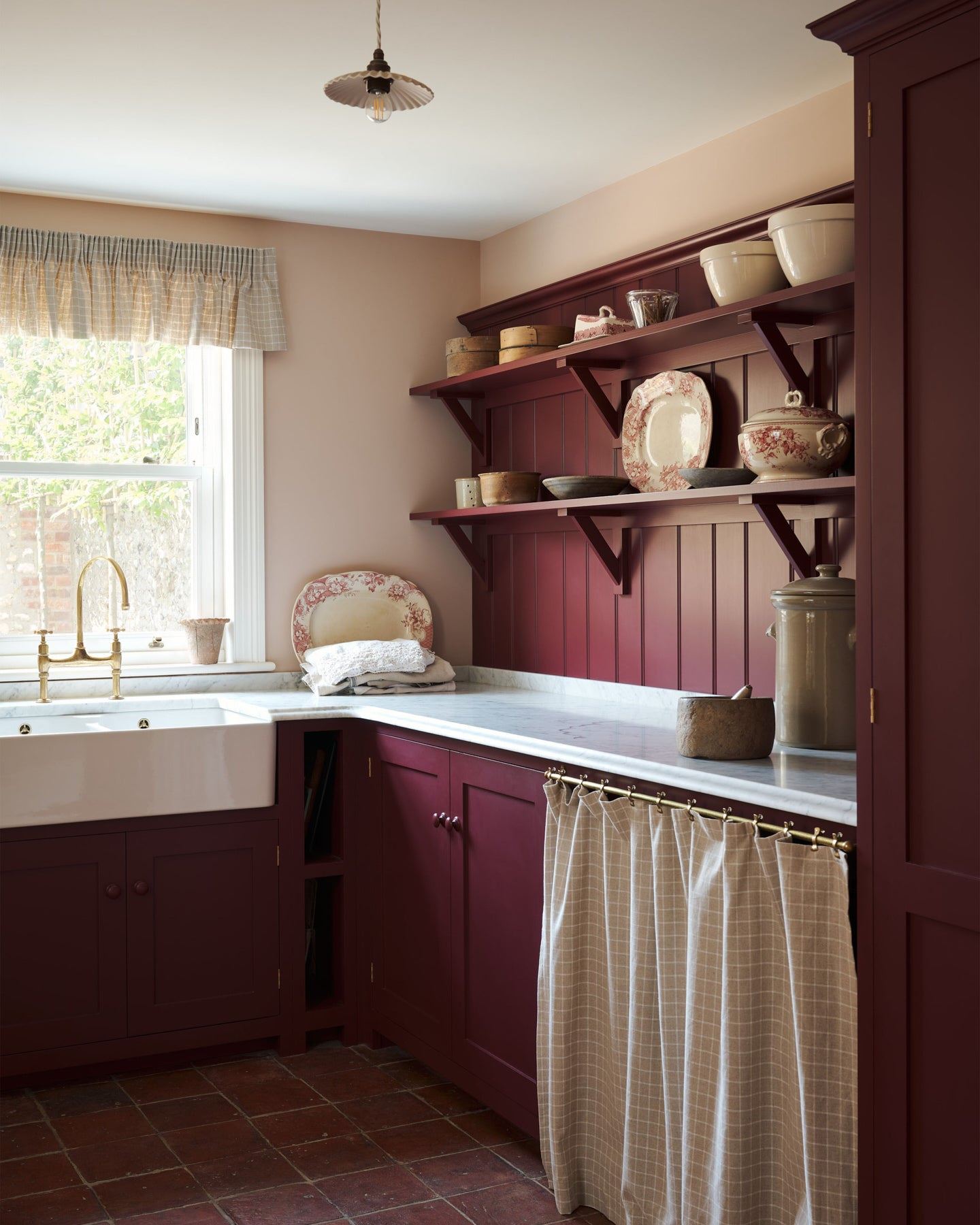 An oblique view of a red painted Shaker-style utility kitchen with a tall pantry cupboard, open storage behind a red striped curtain and tongue and groove wall panelling and shallow shelves decorated with vintage crockery.