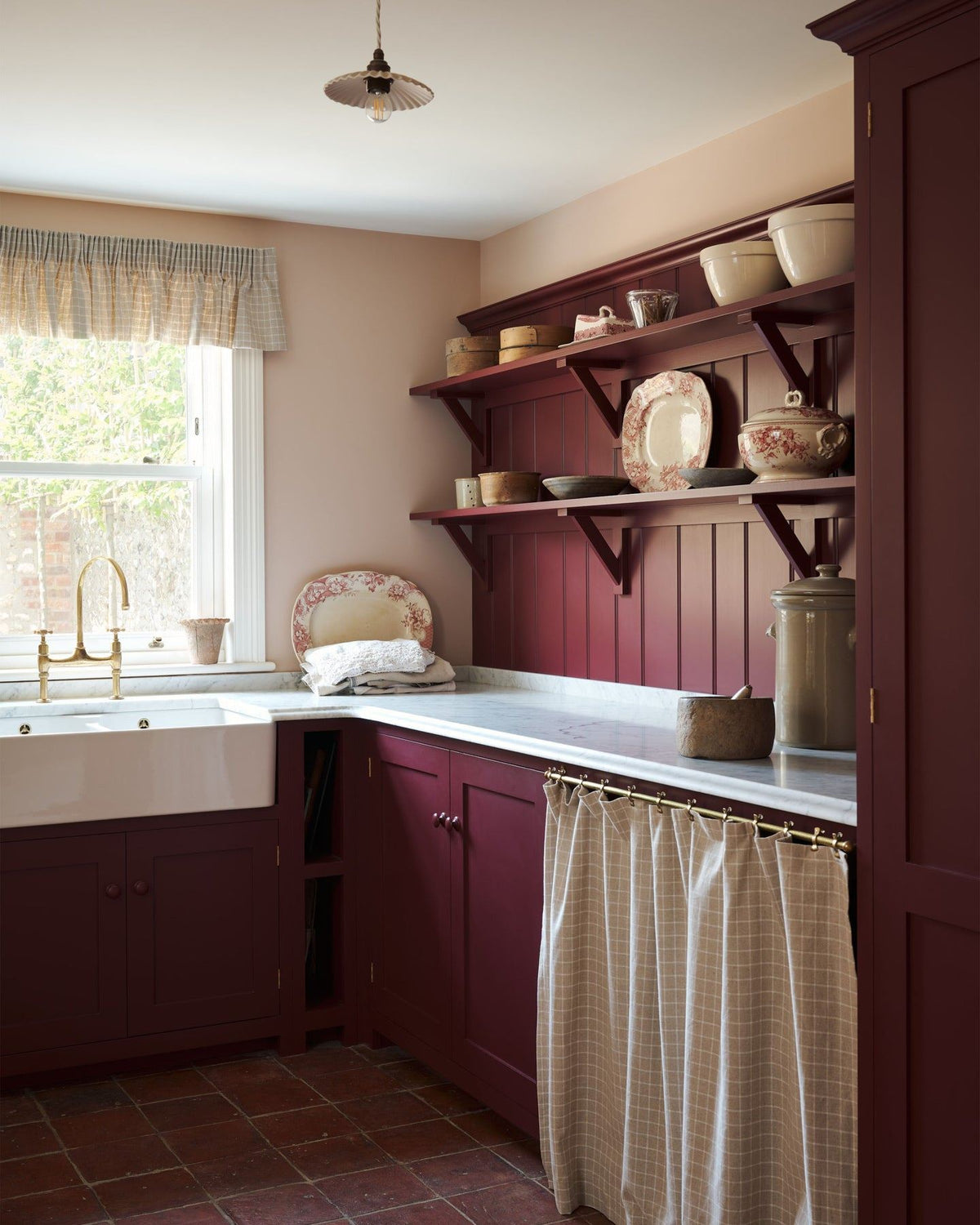 An oblique view of a red painted Shaker-style utility kitchen with a tall pantry cupboard, open storage behind a red striped curtain and tongue and groove wall panelling and shallow shelves decorated with vintage crockery. TEST