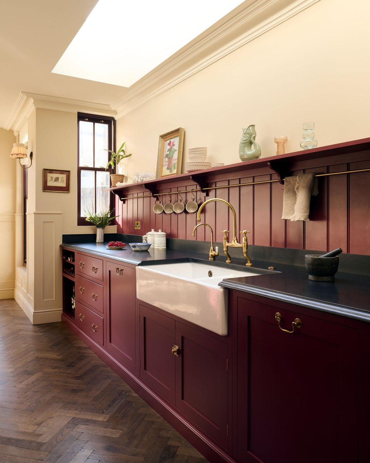 An oblique view of a red painted Shaker-style kitchen with parquet wood flooring, cream walls, a black granite worktop and red tongue and groove panelling above. TEST