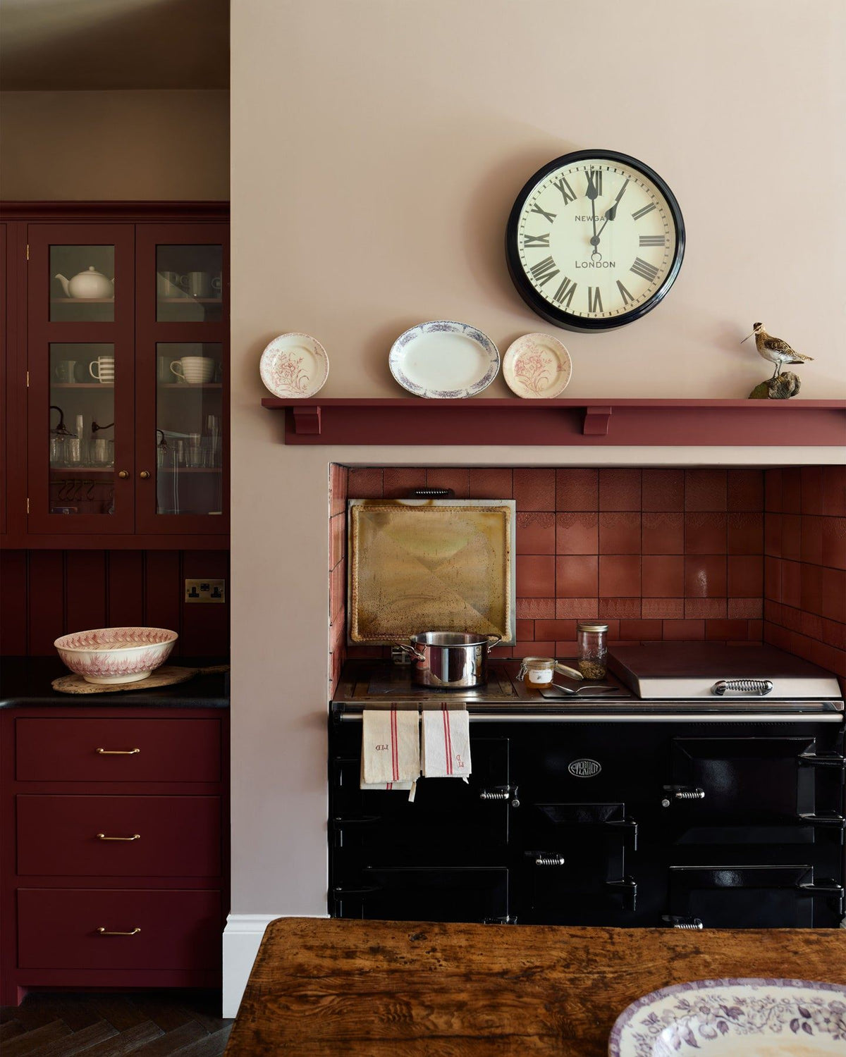 A straight-on view of a red painted Shaker-style kitchen with a large black Aga cooker in a chimney breast alcove with glossy red patterned wall tiles above. TEST