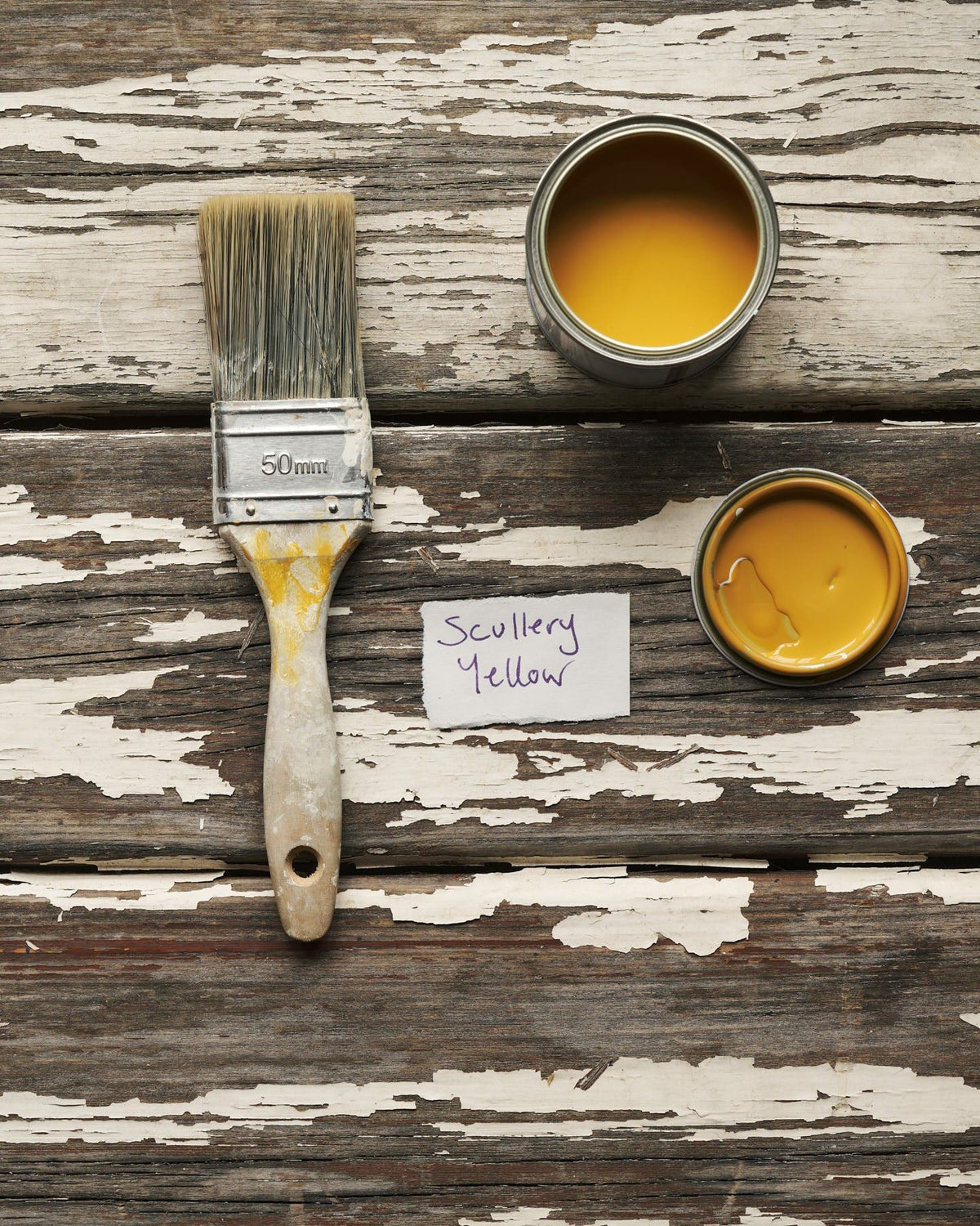 An overhead view of an open paint pot and a worn brush on a rustic tabletop. The warm yellow colour inside is labelled 'Scullery Yellow'. TEST
