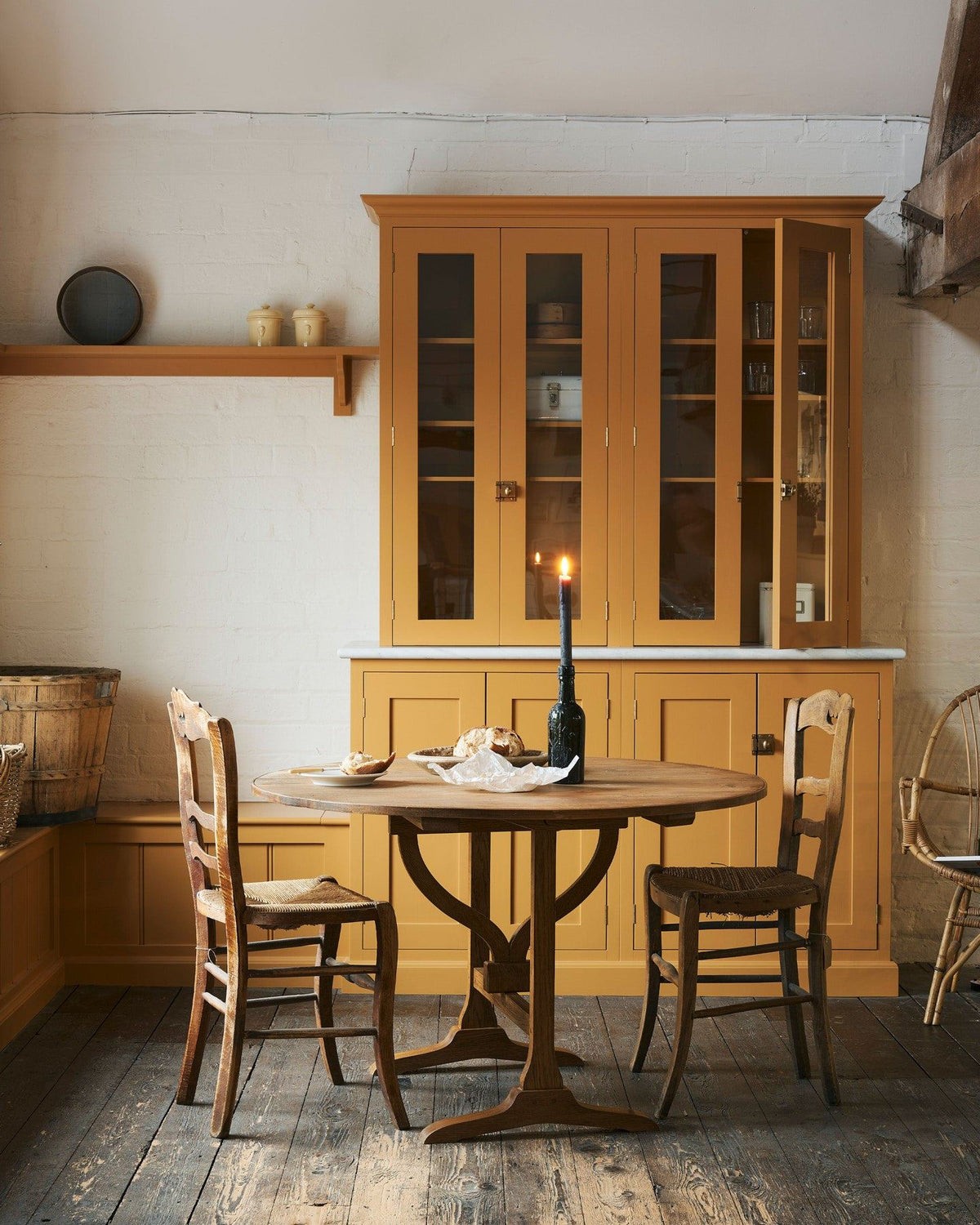A straight-on view of a round pine dining table with vintage wooden chairs, in a rustic cottage kitchen with yellow painted Shaker-style cabinetry, dark worn floorboards, white brick walls and wooden beams. TEST