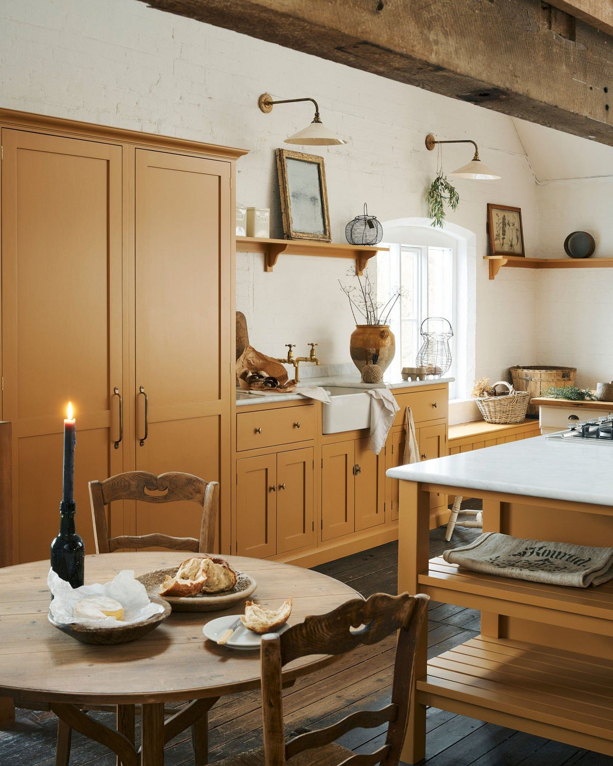 An oblique view of a yellow painted Shaker-style kitchen in a rustic cottage kitchen with dark worn floorboards, white brick walls and wooden beams. TEST