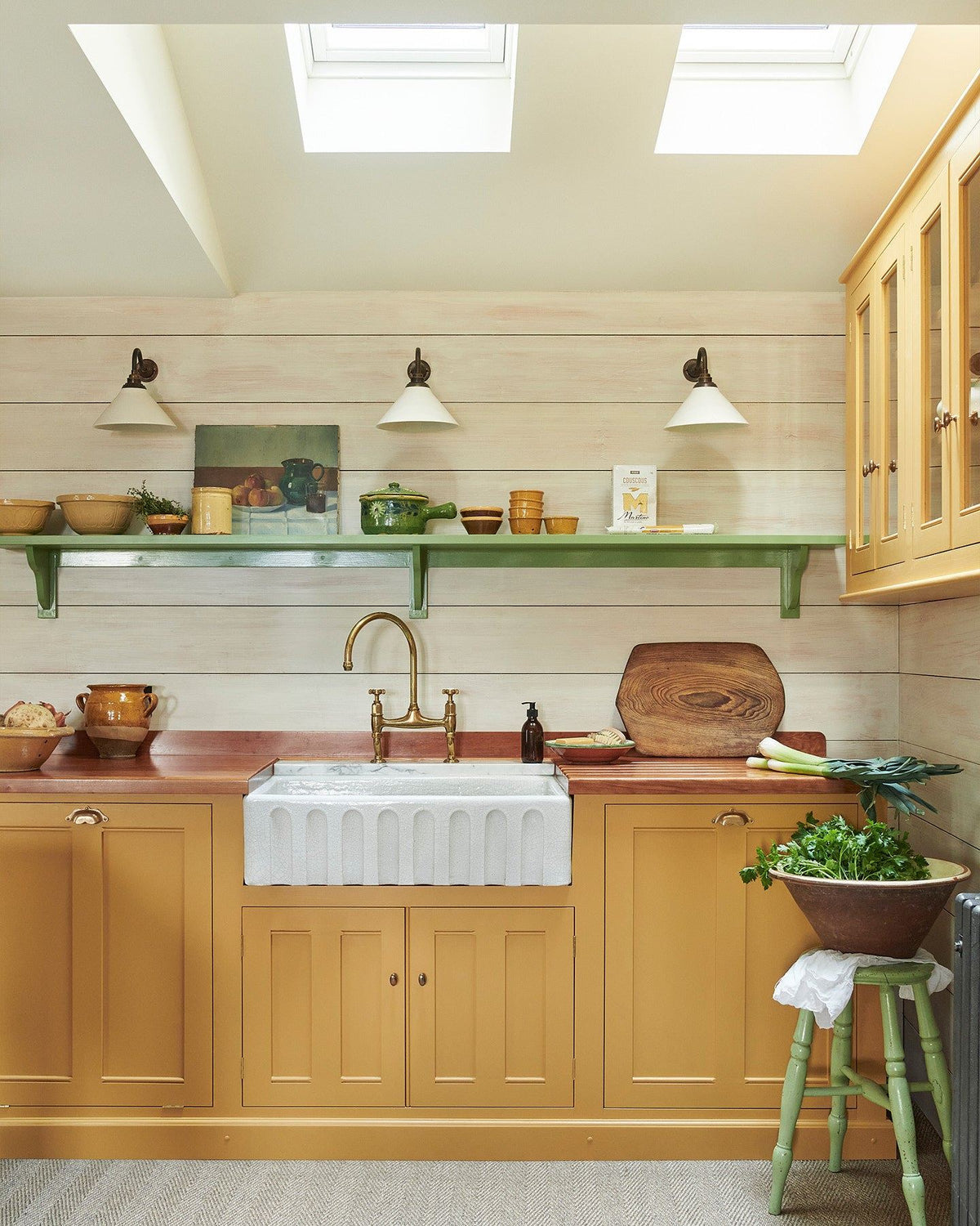 A straight-on view of a yellow painted Georgian-style kitchen sink cabinet in a small cottage kitchen with white painted panelled walls, green shelving and grey carpet flooring. TEST