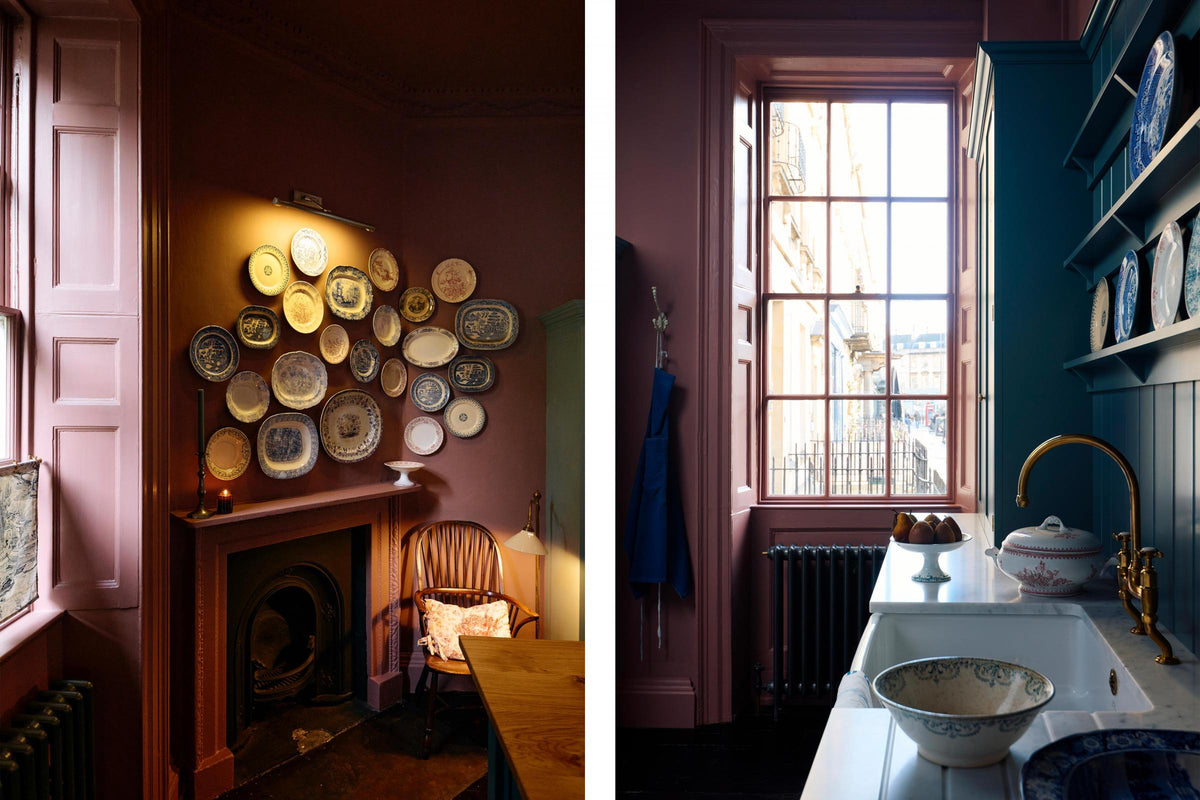 Light streams through sash windows into a moody room with original Georgian features, dusky wine-coloured walls, and dark teal Shaker cabinetry. The display includes a sink run with a fluted ceramic sink, aged brass taps, classic brass door hardware, a white Carrara marble worktop, a tall countertop cupboard, tongue-and-groove wall panelling, and shelves showcasing a selection of classic decorative plates.