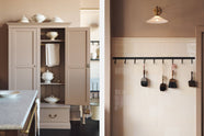 A quiet corner of a grand Princelet pink Heirloom and Classic English kitchen features a sink cabinet with a black granite worktop, a large kitchen island with a Carrara marble worktop, aged brass rails, ditsy delft wallpaper, a tall cream subway tile splashback, and an Heirloom task light. Lovingly adorned with an iron hook rack, antique brushes, an Heirloom task light, and artisan ceramics, creating a warm, delicate, and airy kitchen space.