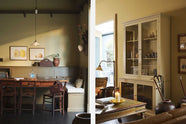 A simple buttery yellow Shaker living space features a Folgate green bespoke bench seat with an open base cabinet, a shelf, a rise and fall pendant light, a curiosity cupboard, and a cohesive set of vintage table and chairs. Lovingly adorned with artisan ceramics, paintings, books, a coat rack and cushions, creating a cosy and Mediterranean cafe-inspired social space.