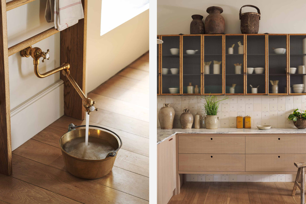 An all-natural and functional Sebastian Cox kitchen features an L-shaped sink run with a marble worktop, glazed wall cupboards, and an aged brass filler tap with a brass pan for a makeshift dog bowl. Lovingly adorned with antique artisan pottery, ceramic pendant lights, beautiful houseplants, and beloved figurines, it creates a wonderfully simple kitchen with a love of natural sustainability.