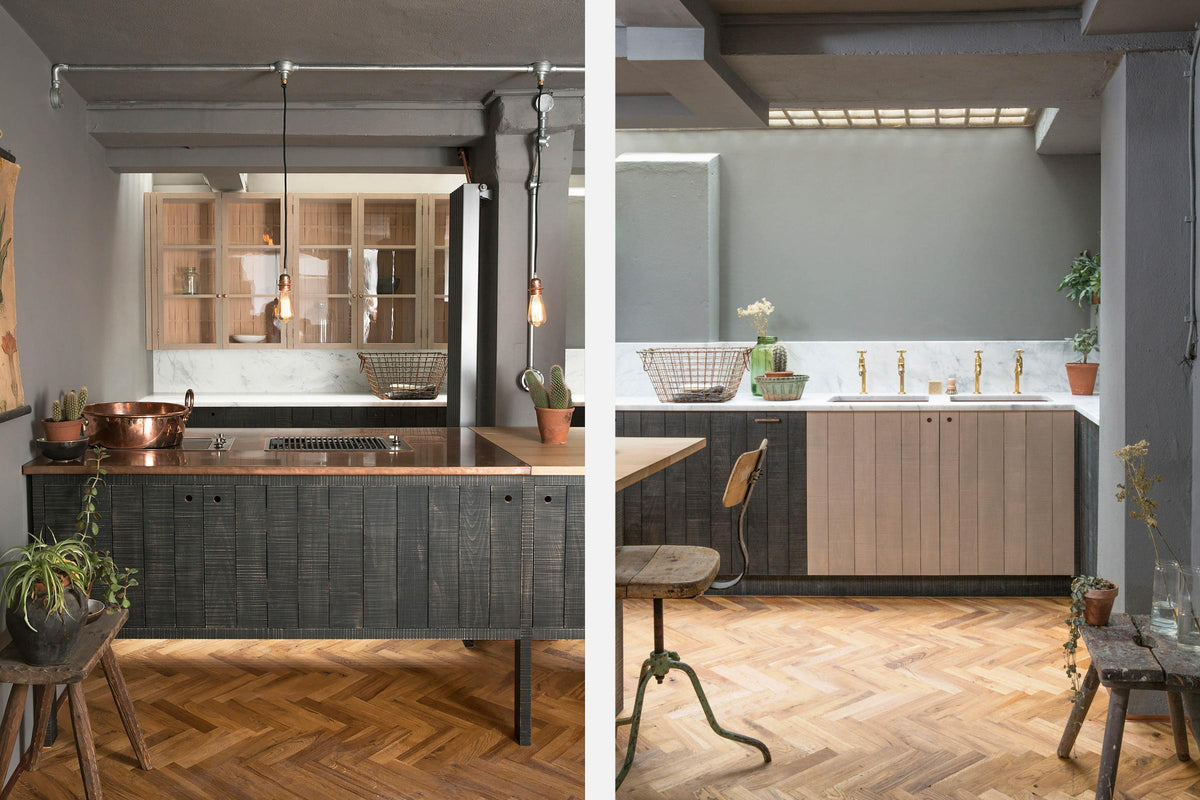 A chic and simple Sebastian Cox basement kitchen features a long sink run with two sets of copper sinks, aged brass Mayan taps, a Carrara marble worktop, glazed wall cupboards, a large kitchen island with a gas cooker, a copper worktop, and a breakfast nook. This kitchen creates a cool and atmospheric space, beautifully decorated with luscious green house plants, vintage items, drinking glasses, a plumber’s stool, and a simple pendant light.