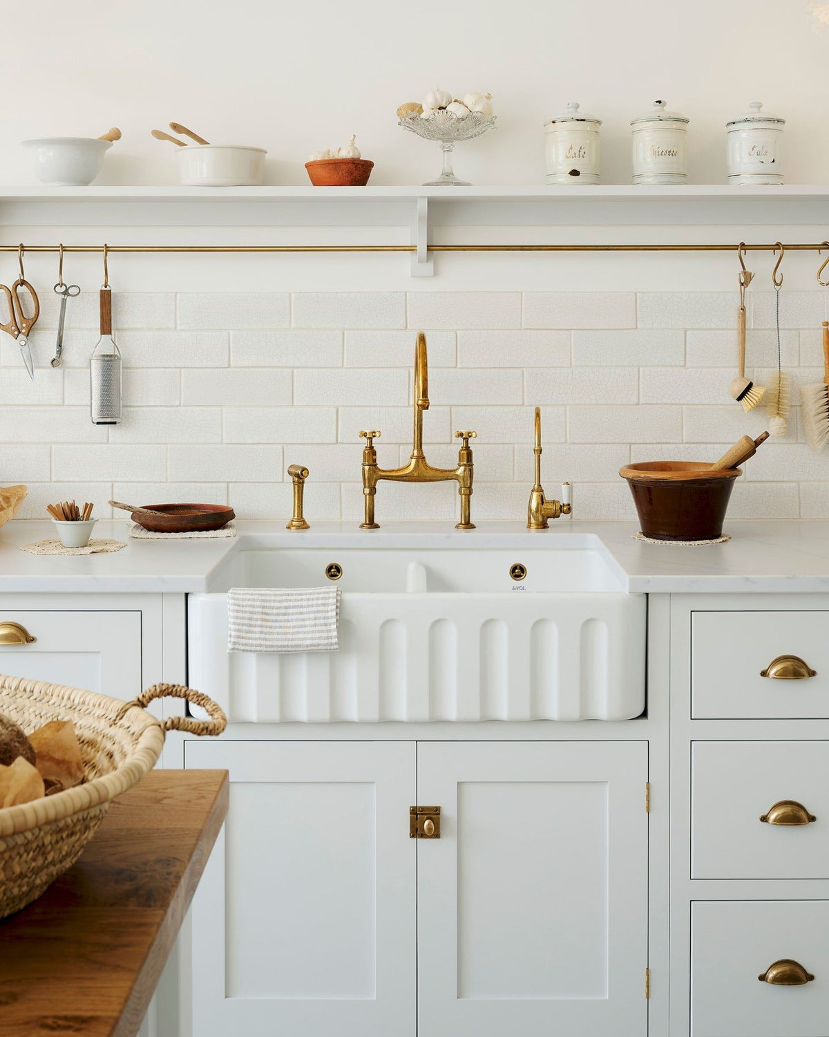 A straight-on view of a large fluted double bowl ceramic Belfast sink in a white painted simple Shaker-style kitchen with an aged brass mixer tap and white metro wall tiles. TEST