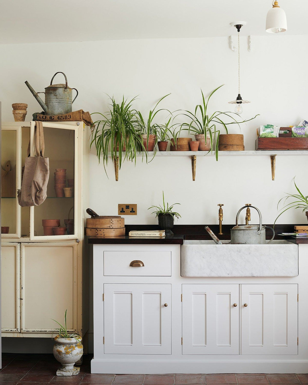 A straight-on view of a classic 13 amp twin plug socket in a mottled aged brass finish in a Mediterannean-inspired kitchen with a white painted Georgian-style sink cabinet, dark hardwood worktop, Carrara marble sink and a shelf full of lush spider plants. TEST