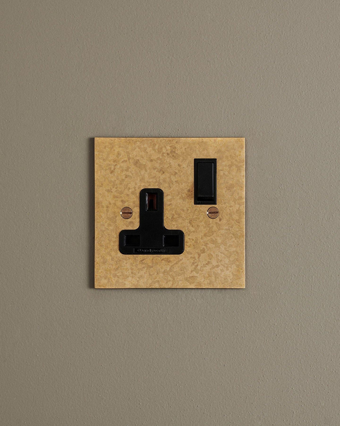 A straight-on view of a classic 13 amp single plug socket in a mottled aged brass finish with black plastic switches.