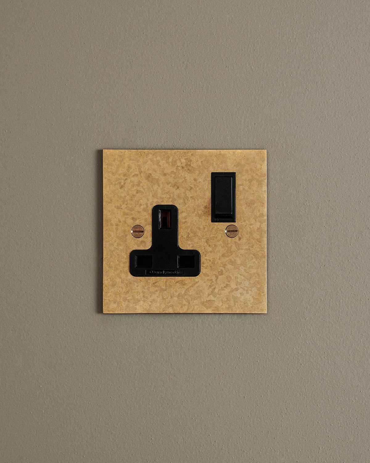 A straight-on view of a classic 13 amp single plug socket in a mottled aged brass finish with black plastic switches. TEST