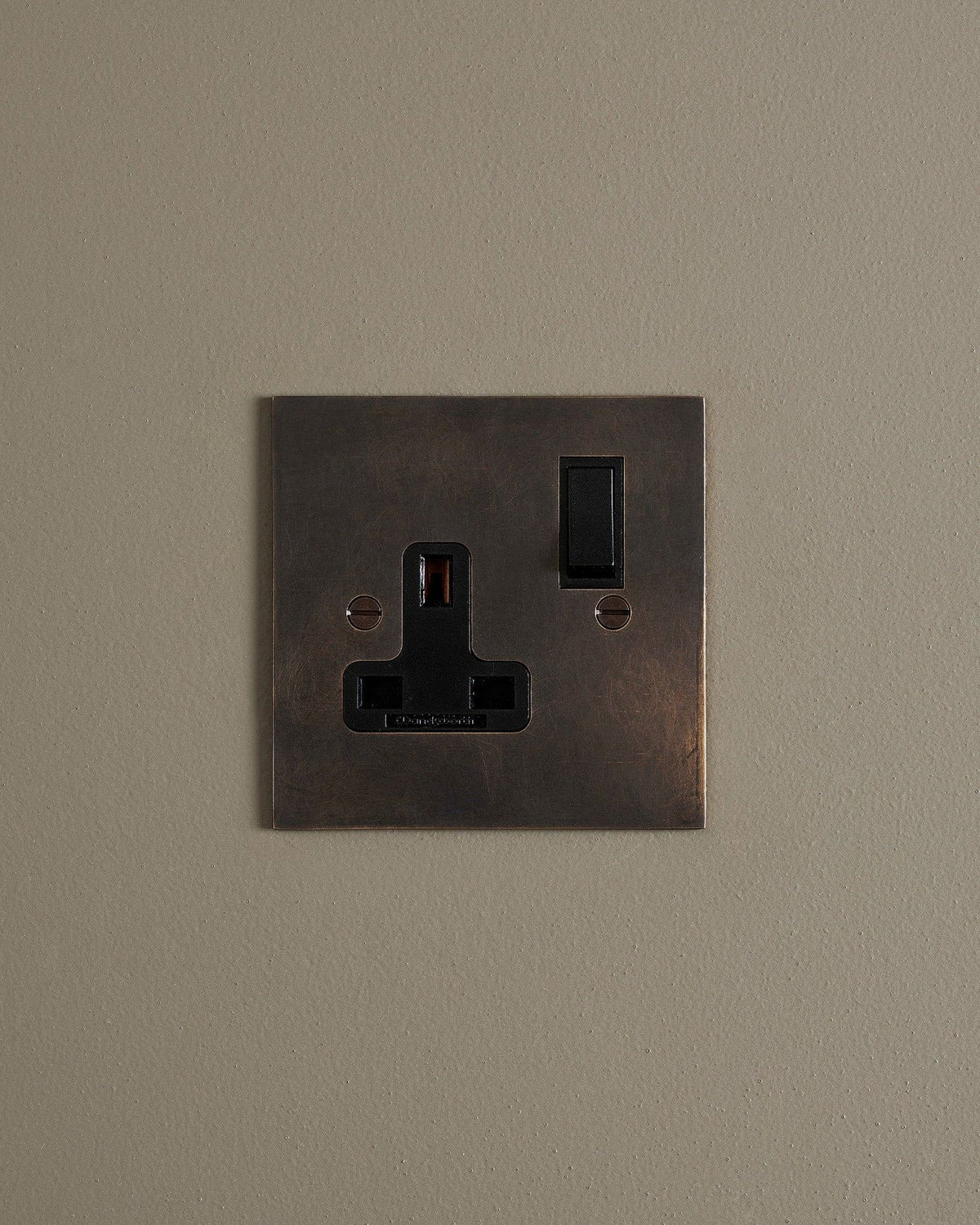 A straight-on view of a classic 13 amp single plug socket in a dark oxidised brass finish with black plastic switches. TEST
