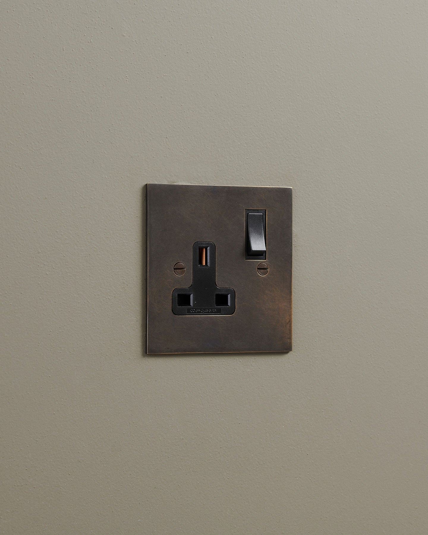 An oblique view of a classic 13 amp single plug socket in a dark oxidised brass finish with black plastic switches.