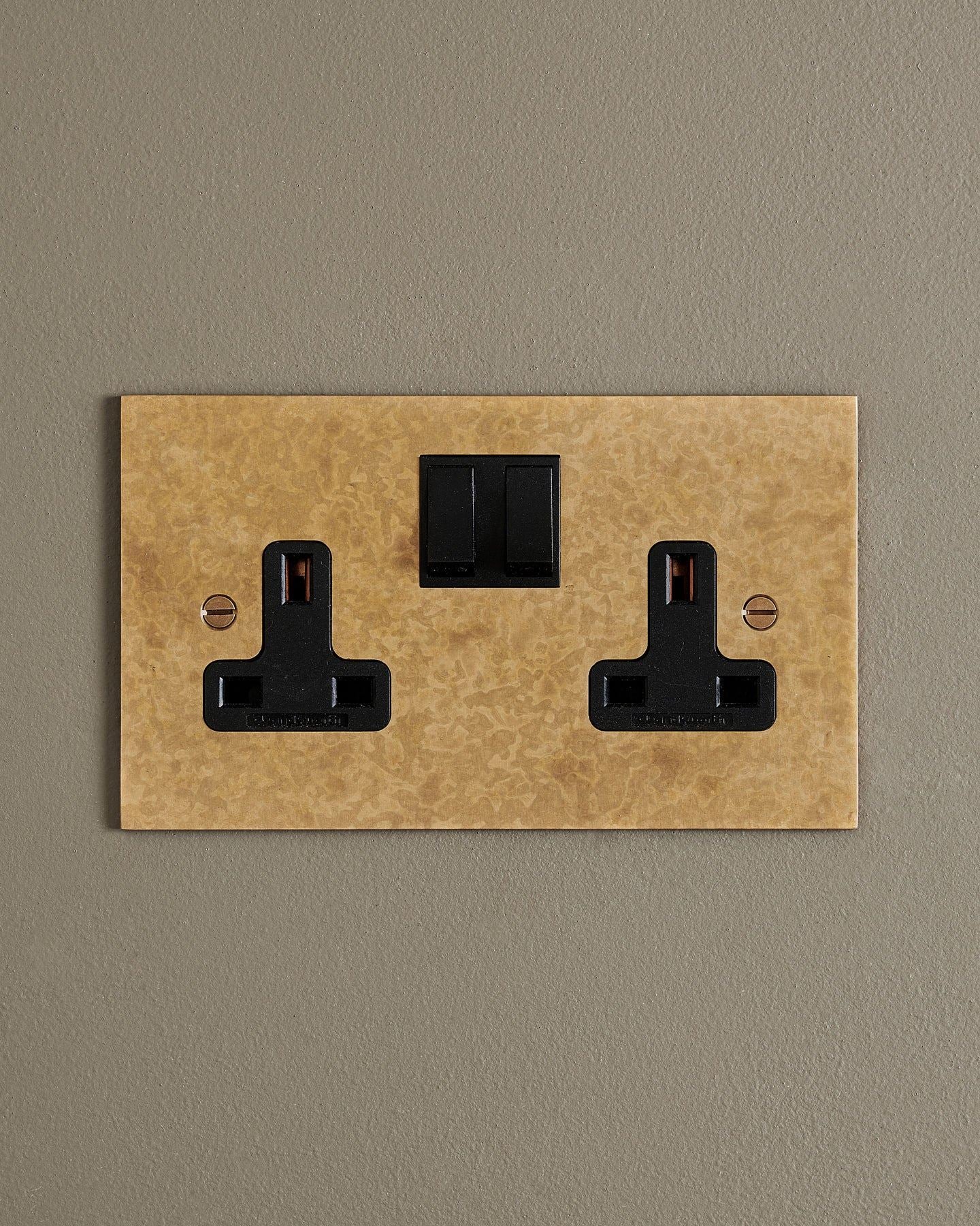 A straight-on view of a classic 13 amp twin plug socket in a mottled aged brass finish with black plastic switches.