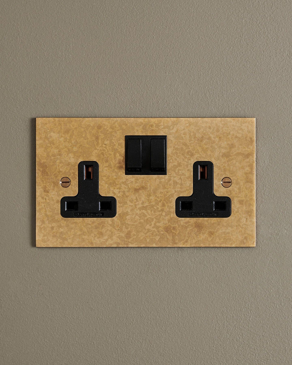 A straight-on view of a classic 13 amp twin plug socket in a mottled aged brass finish with black plastic switches. TEST