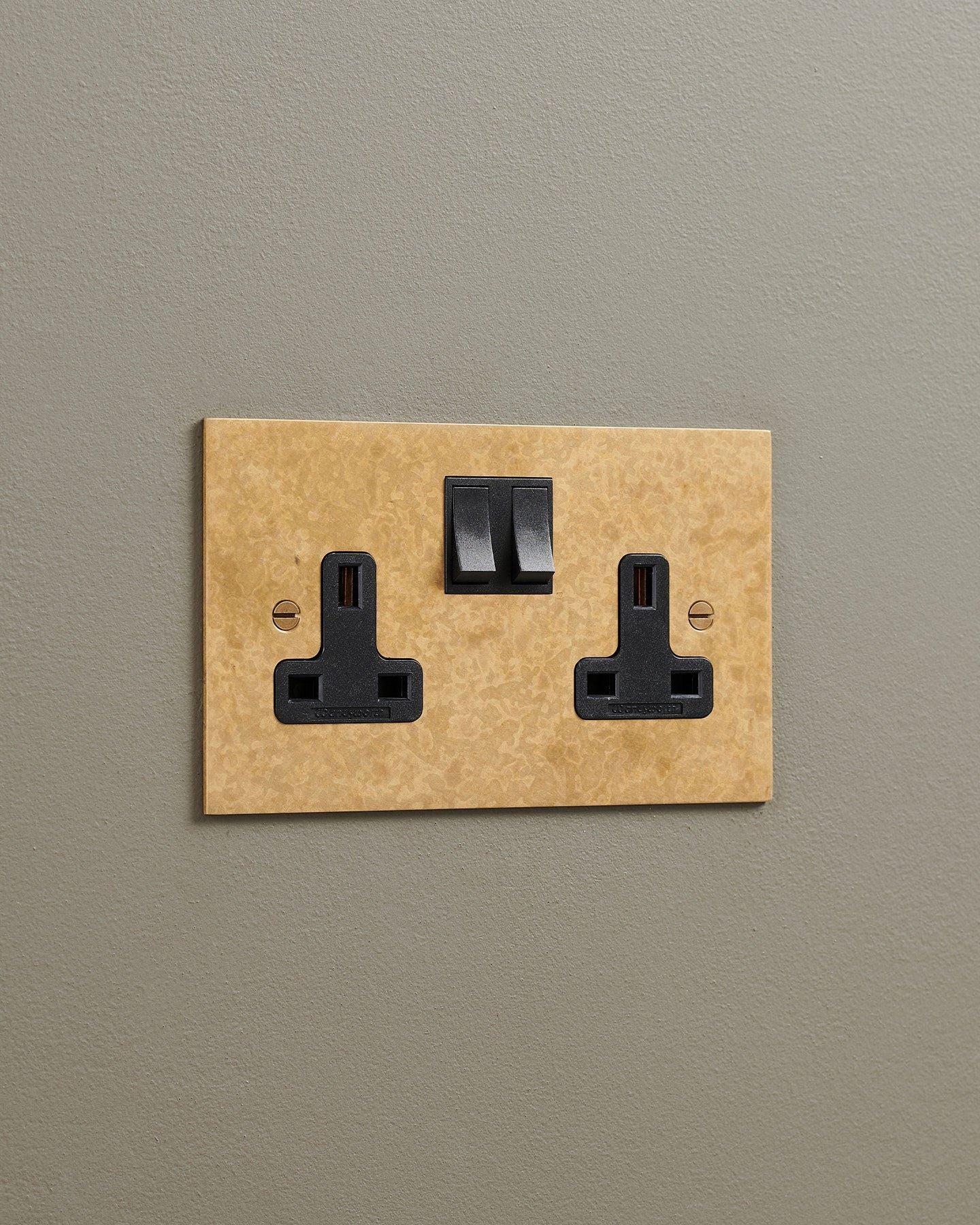 An oblique view of a classic 13 amp twin plug socket in a mottled aged brass finish with black plastic switches.