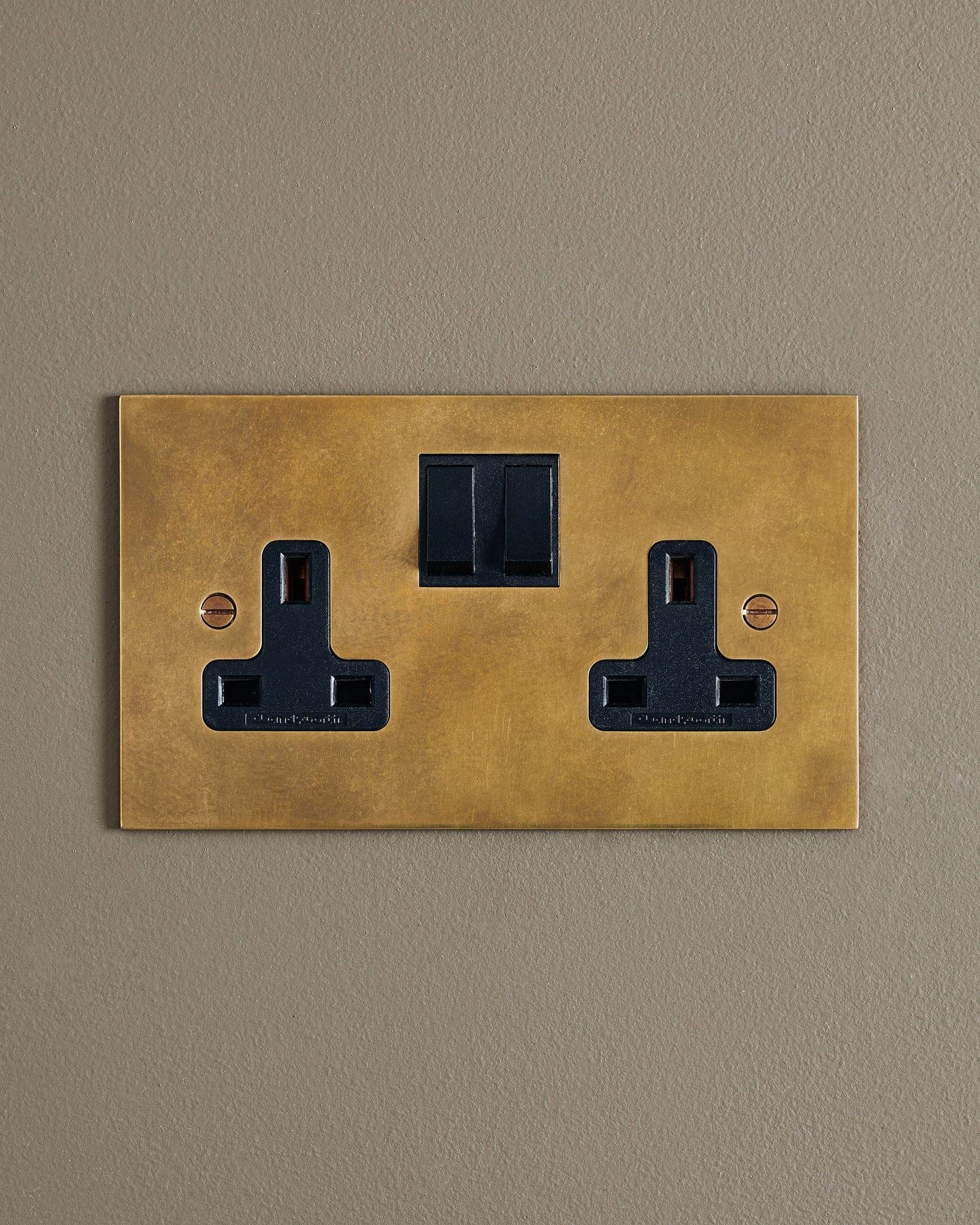 A straight-on view of a classic 13 amp twin plug socket in a warm burnished brass finish with black plastic switches.