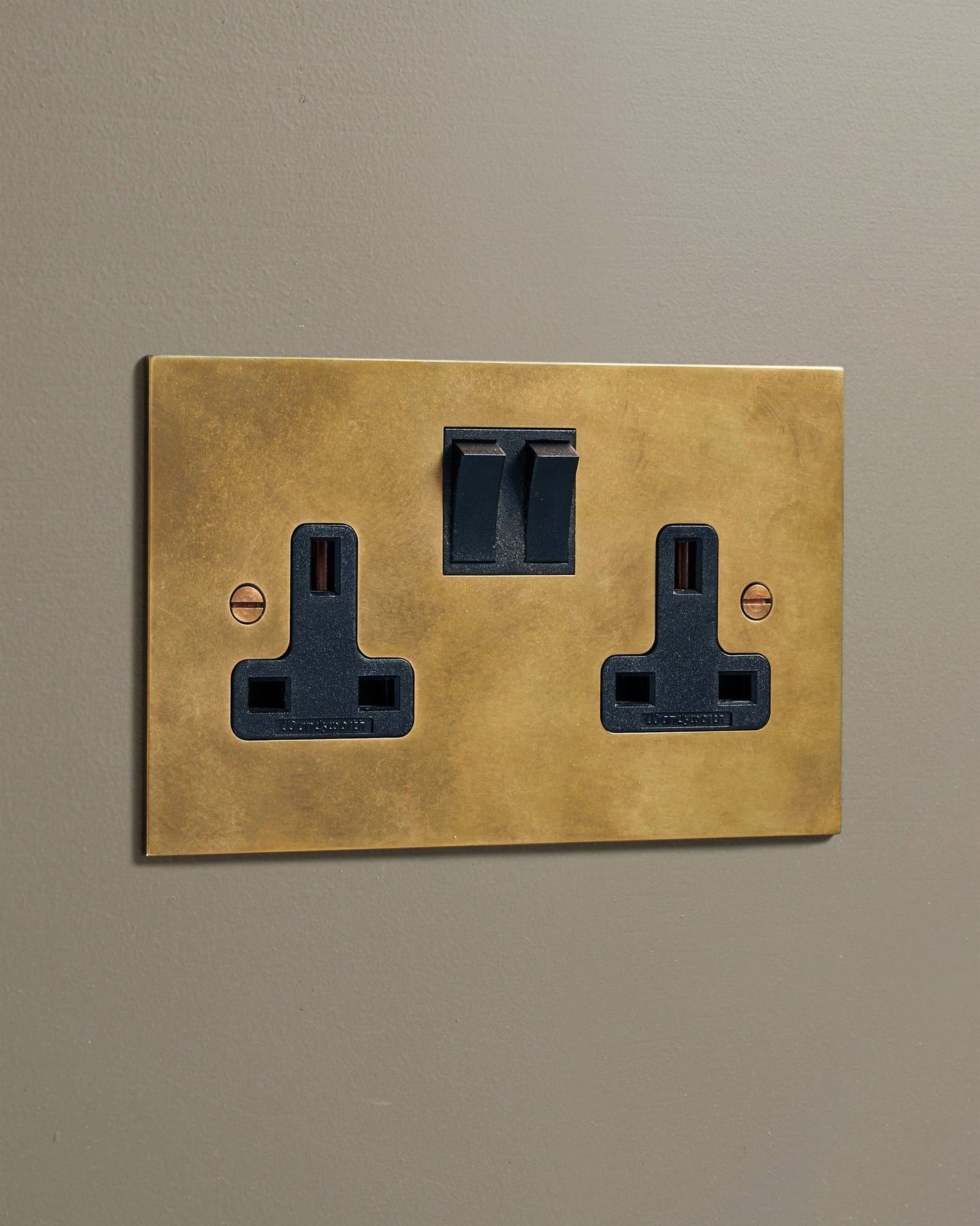 An oblique view of a classic 13 amp twin plug socket in a warm burnished brass finish with black plastic switches.
