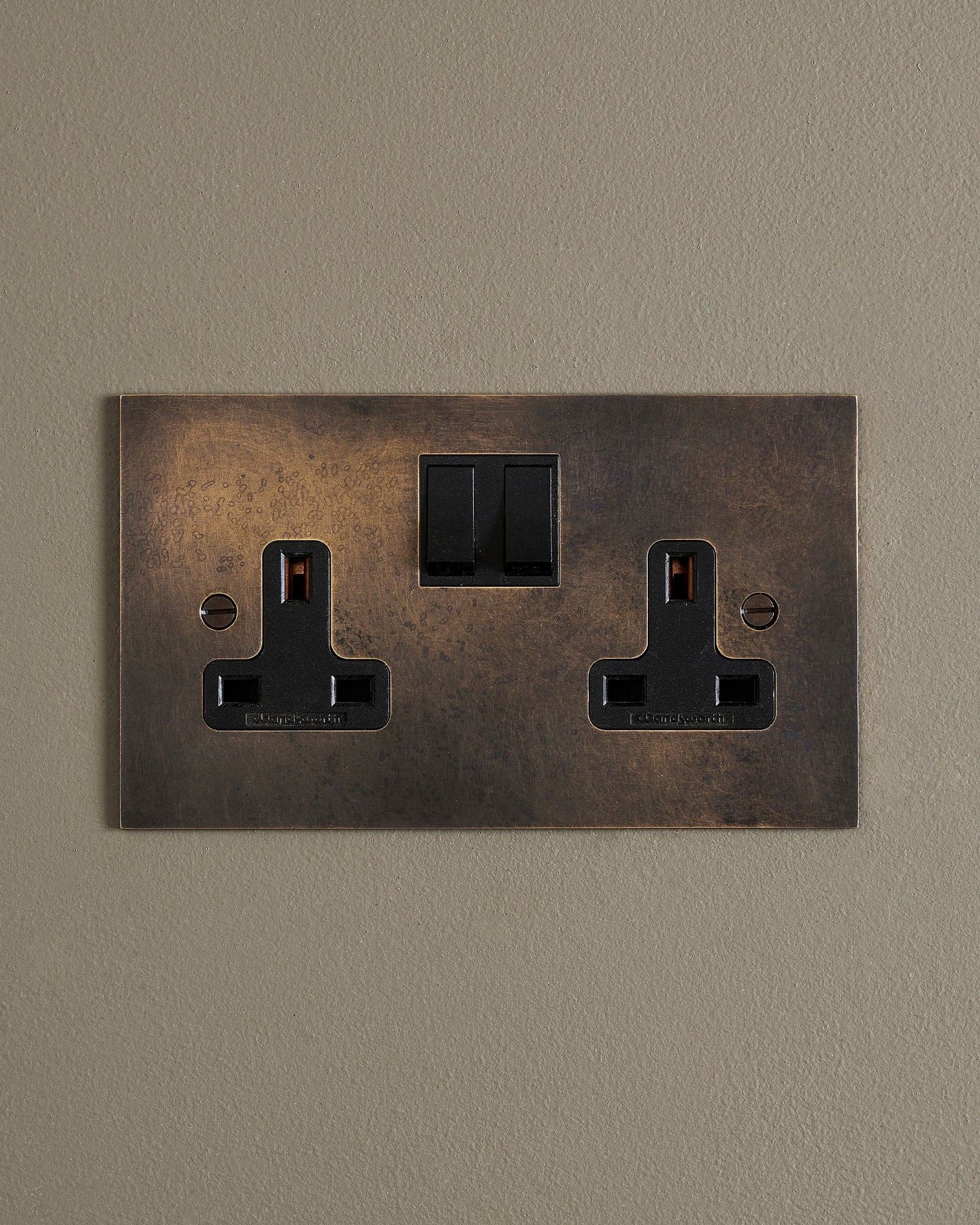 A straight-on view of a classic 13 amp twin plug socket in a dark oxidised brass finish with black plastic switches.
