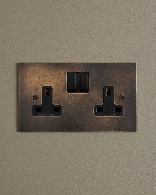 A straight-on view of a classic 13 amp twin plug socket in a dark oxidised brass finish with black plastic switches.