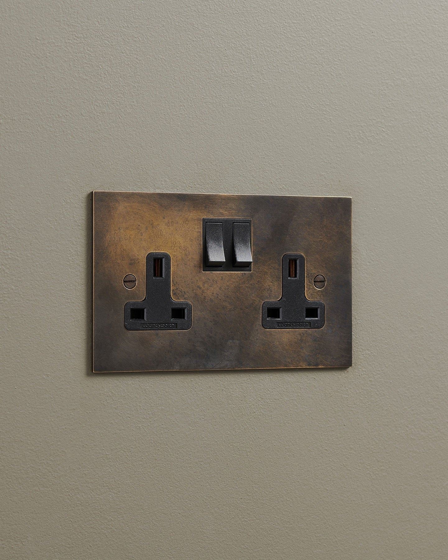 An oblique view of a classic 13 amp twin plug socket in a dark oxidised brass finish with black plastic switches.