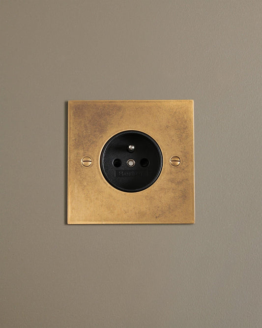 A straight-on view of a classic French-style single plug socket in a mottled aged brass finish.