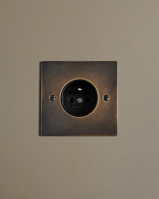 A straight-on view of a classic French-style single plug socket in a dark oxidised brass finish.