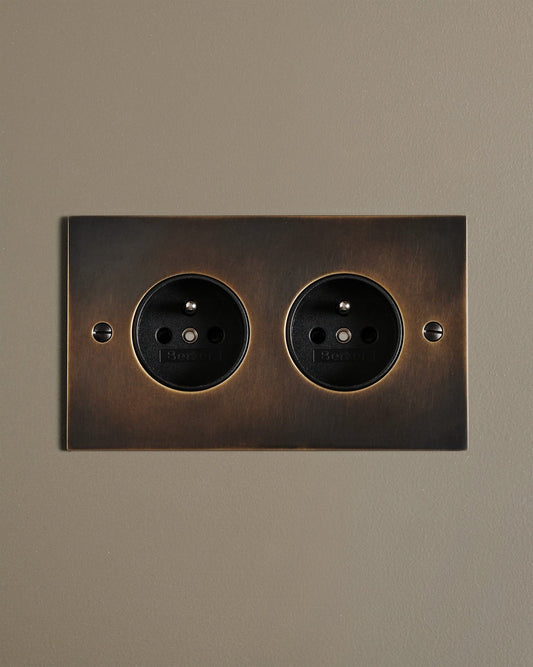 A straight-on view of a classic French-style twin plug socket in a dark oxidised brass finish.