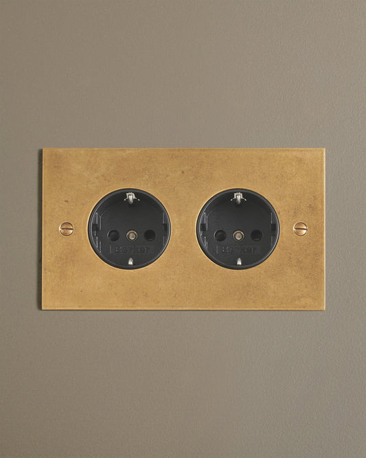 A straight-on view of a classic Schuko-style twin plug socket in a mottled aged brass finish.