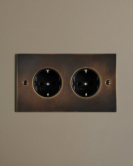 A straight-on view of a classic Schuko-style twin plug socket in a dark oxidised brass finish.