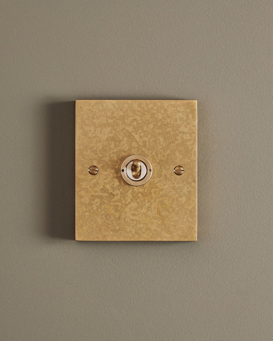 A straight-on view of a 1 gang box toggle switch in a mottled aged brass finish.