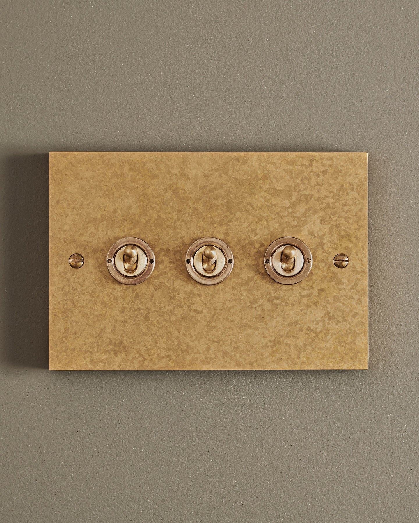 A straight-on view of a 3 gang box toggle switch in a mottled aged brass finish.