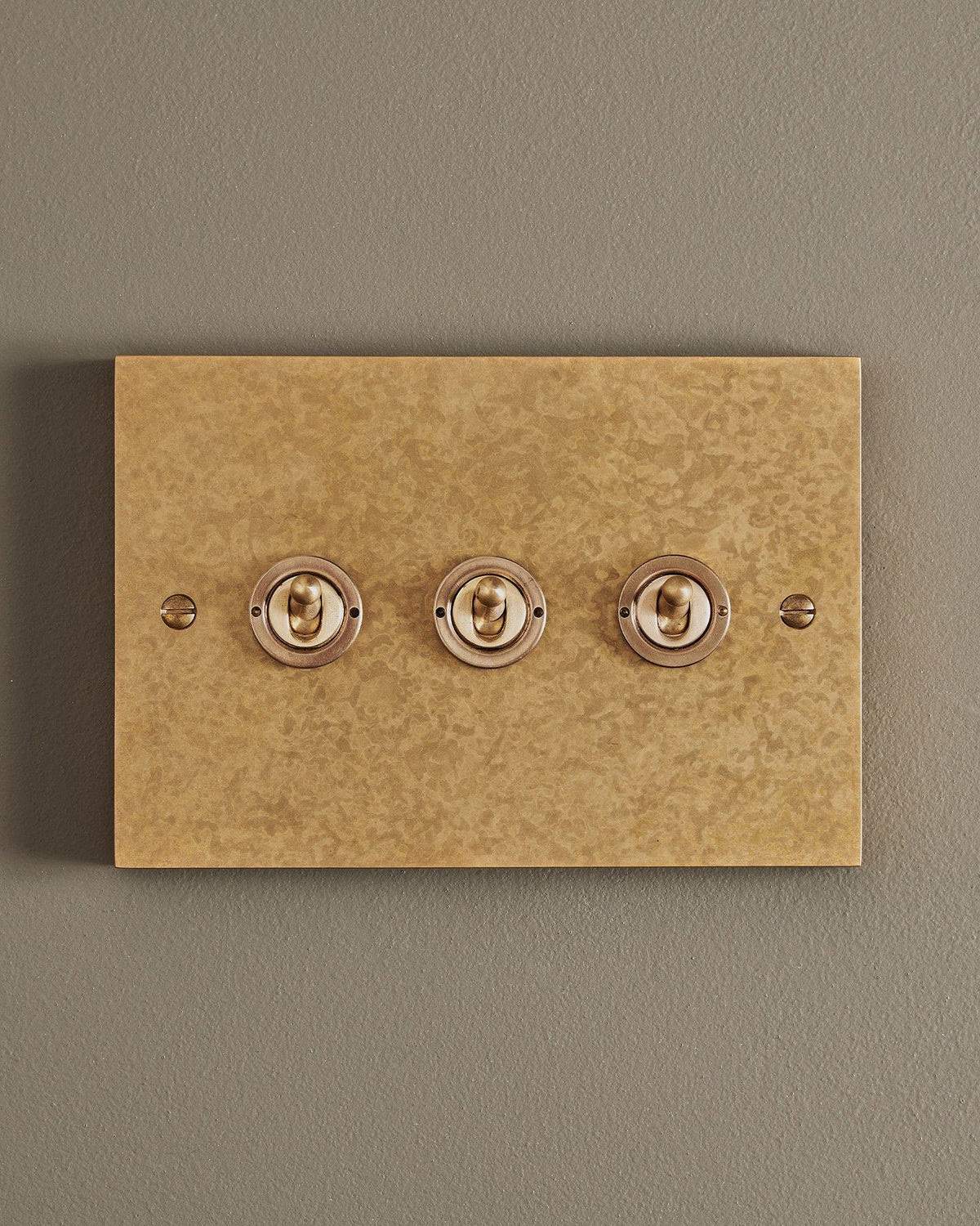 A straight-on view of a 3 gang box toggle switch in a mottled aged brass finish. TEST