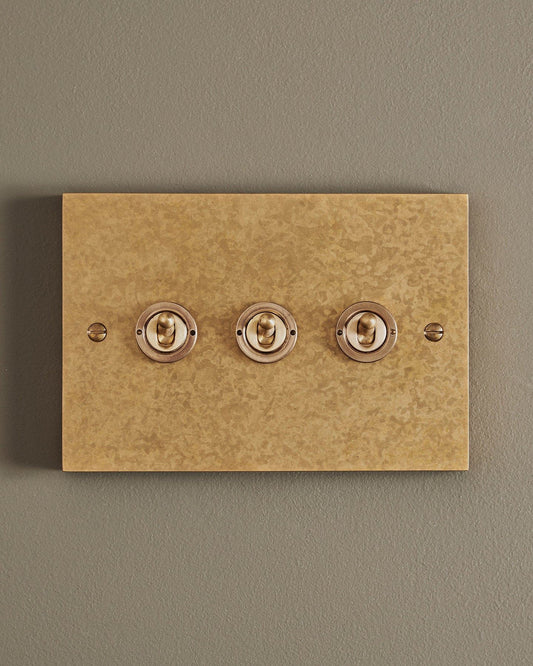 A straight-on view of a 3 gang box toggle switch in a mottled aged brass finish.