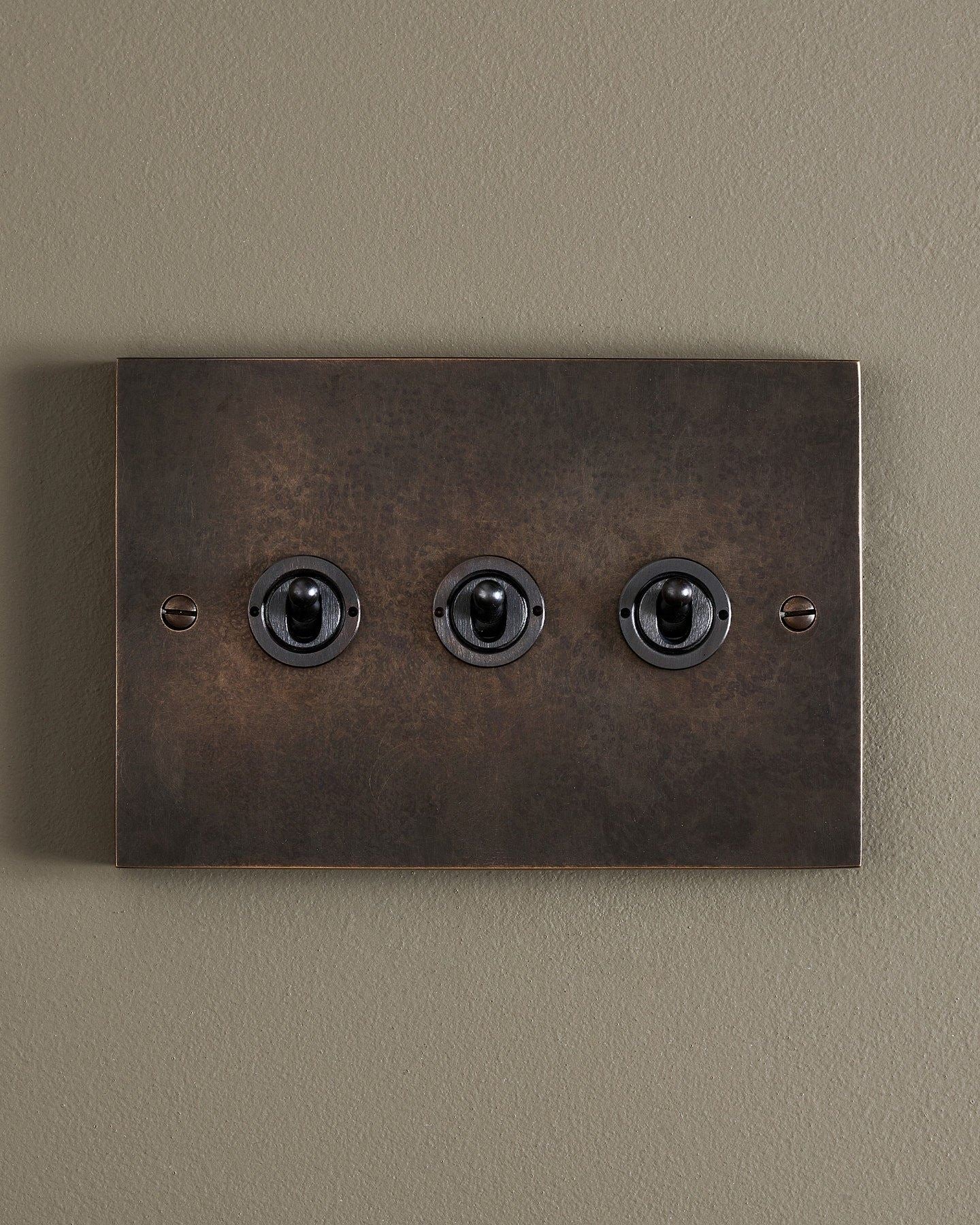 A straight-on view of a 3 gang box toggle switch in a dark oxidised brass finish.