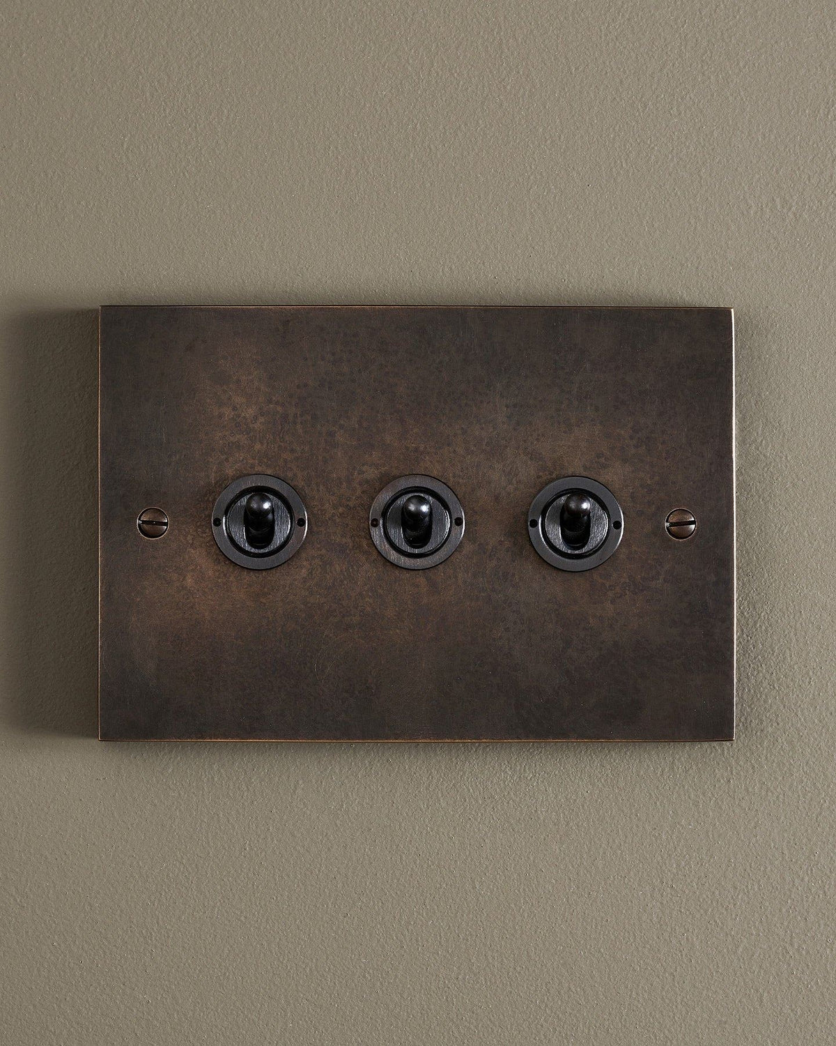 A straight-on view of a 3 gang box toggle switch in a dark oxidised brass finish. TEST