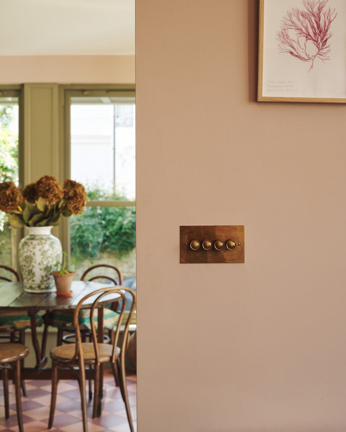 A straight-on view of a 4 gang classic dimmer switch in a warm burnished brass finish on soft pink walls, a cafÈ-style table and chairs with a vase full of pink hydrangeas can be seen in the background.|Heirloom Brass shown TEST