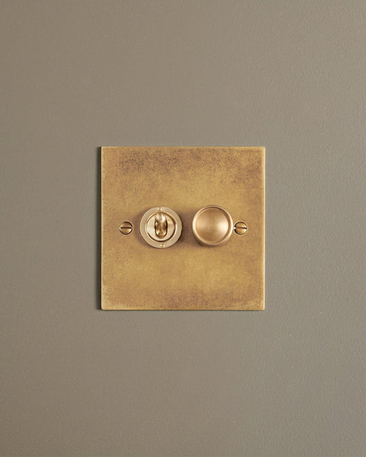 A straight-on view of a 2 gang classic toggle and dimmer mixed switch in a mottled aged brass finish.