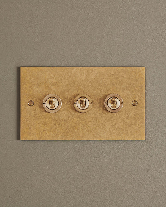 A straight-on view of a 3 gang classic toggle switch in a mottled aged brass finish.