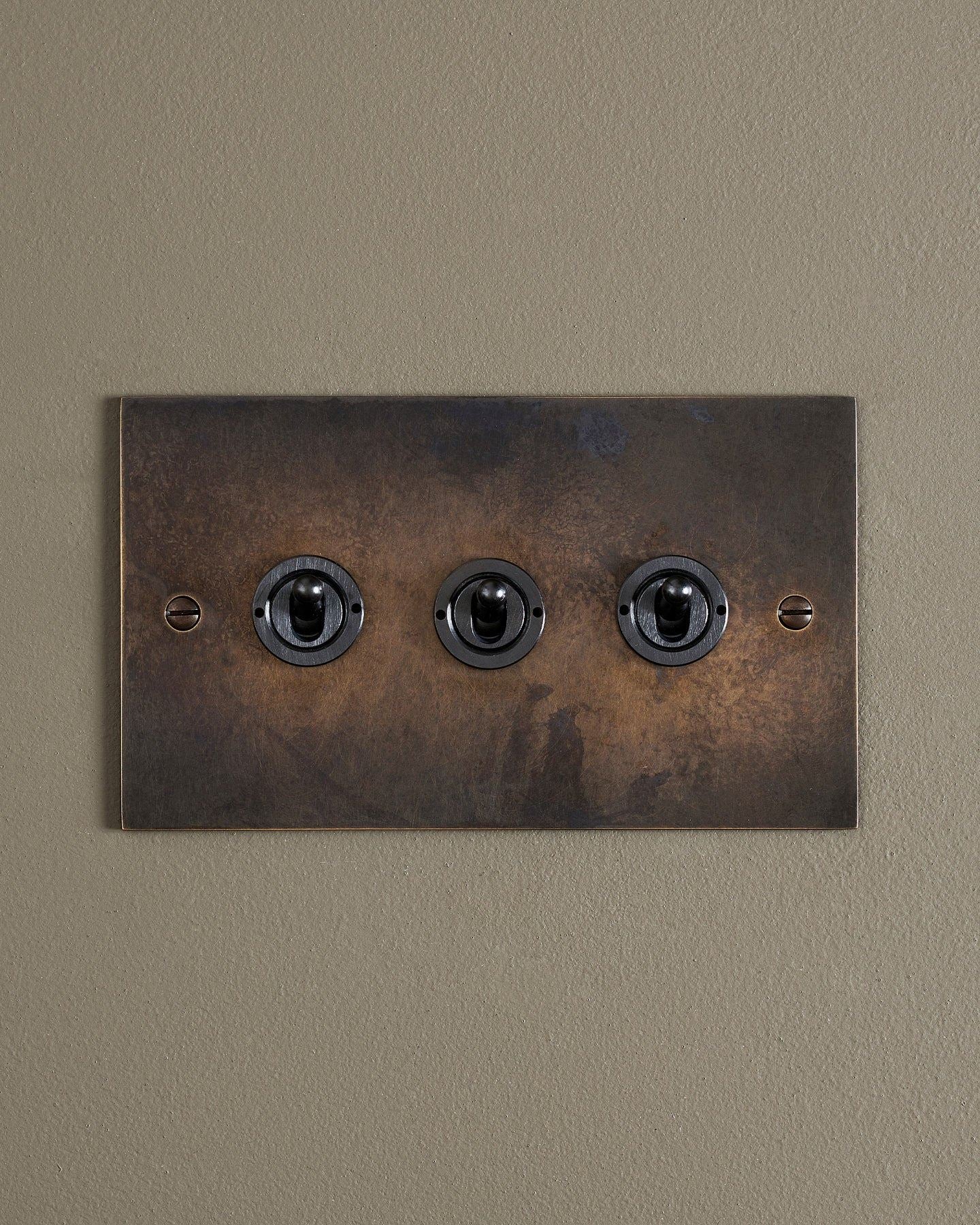 A straight-on view of a 3 gang classic toggle switch in a dark oxidised brass finish.