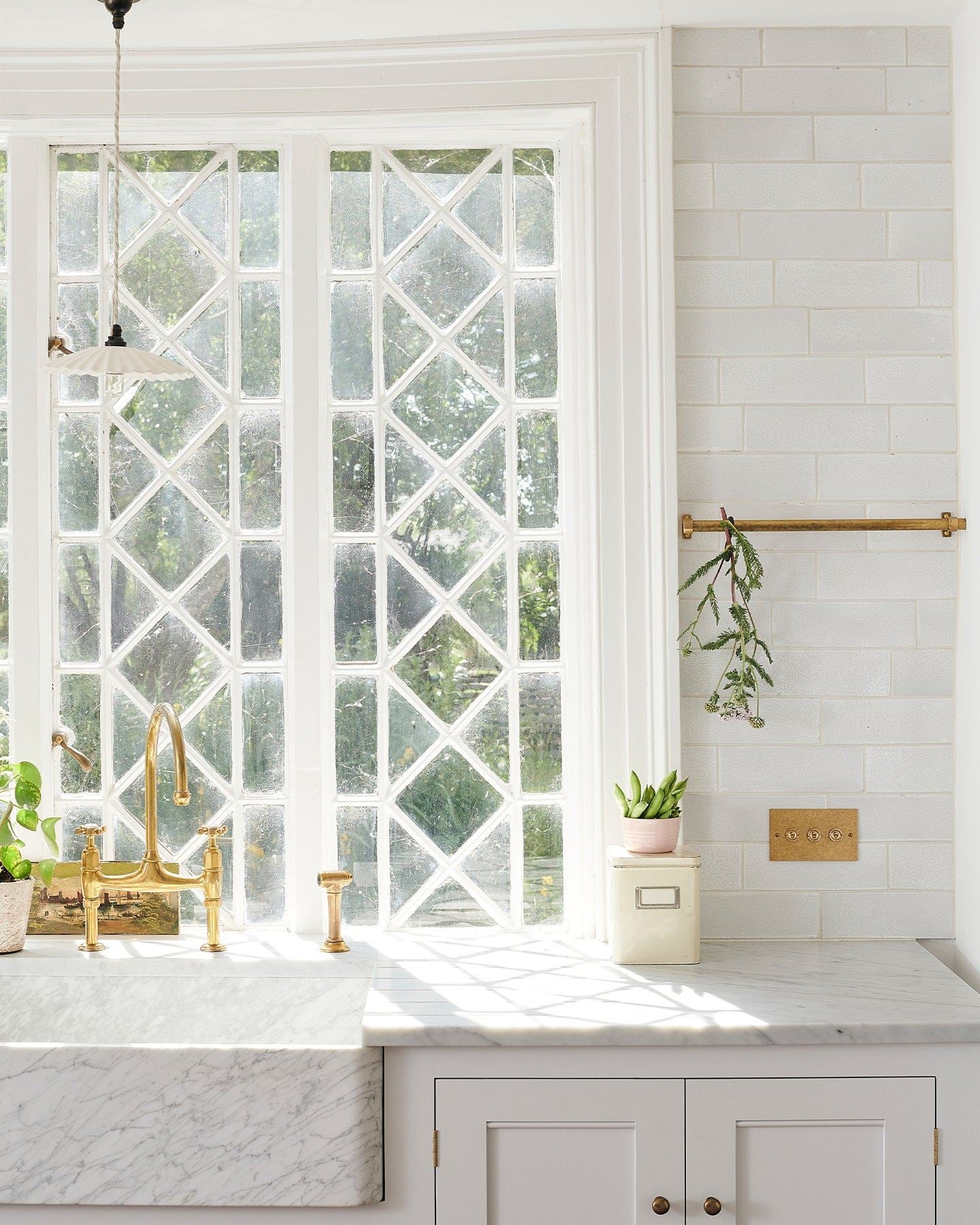 A straight-on view of a 3 gang classic toggle switch in an aged brass finish in a Mediterranean-style kitchen with white painted cupboards, Carrara marble worktop, white metro tiles and original Victorian white leaded windows.
