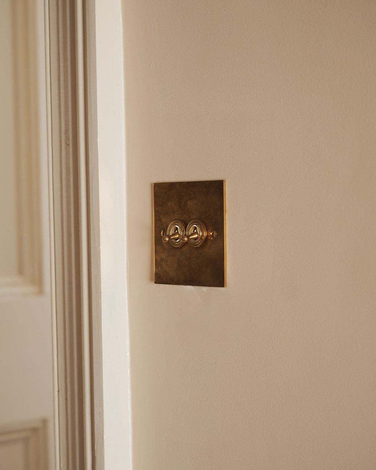 An oblique view of a 2 gang classic toggle switch in a warm burnished brass finish on soft pink walls with light painted woodwork. TEST