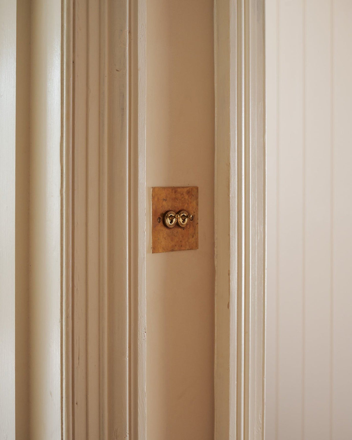 An oblique view of a 2 gang classic toggle switch in a warm burnished brass finish on soft pink walls between traditional light painted architrave. | 2 Gang shown TEST