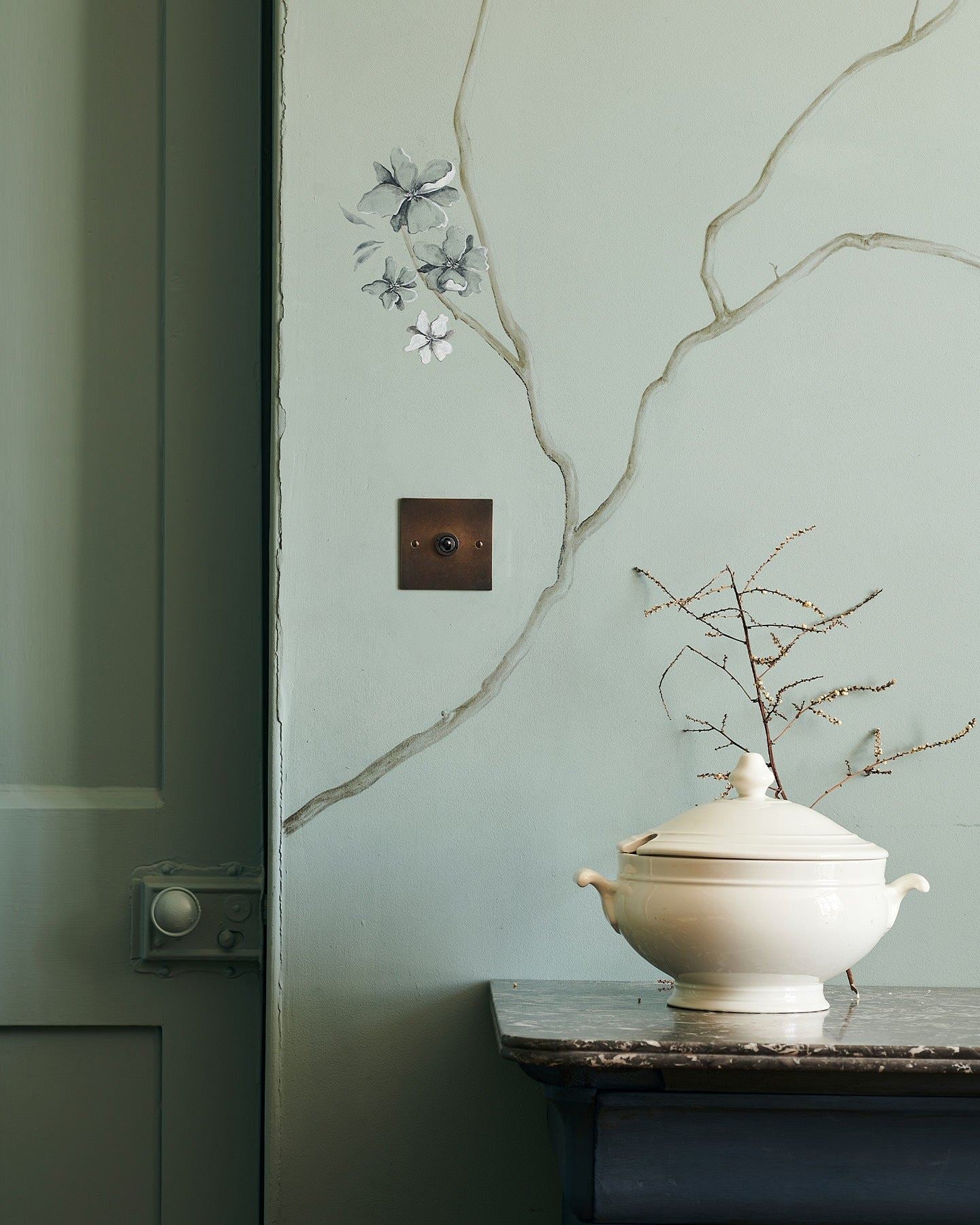 A straight-on view of a 1 gang classic toggle switch in a dark oxidised brass finish on a duck-egg blue wall with a delicate handpainted mural of winding branches and a glossy white pot in the foreground.