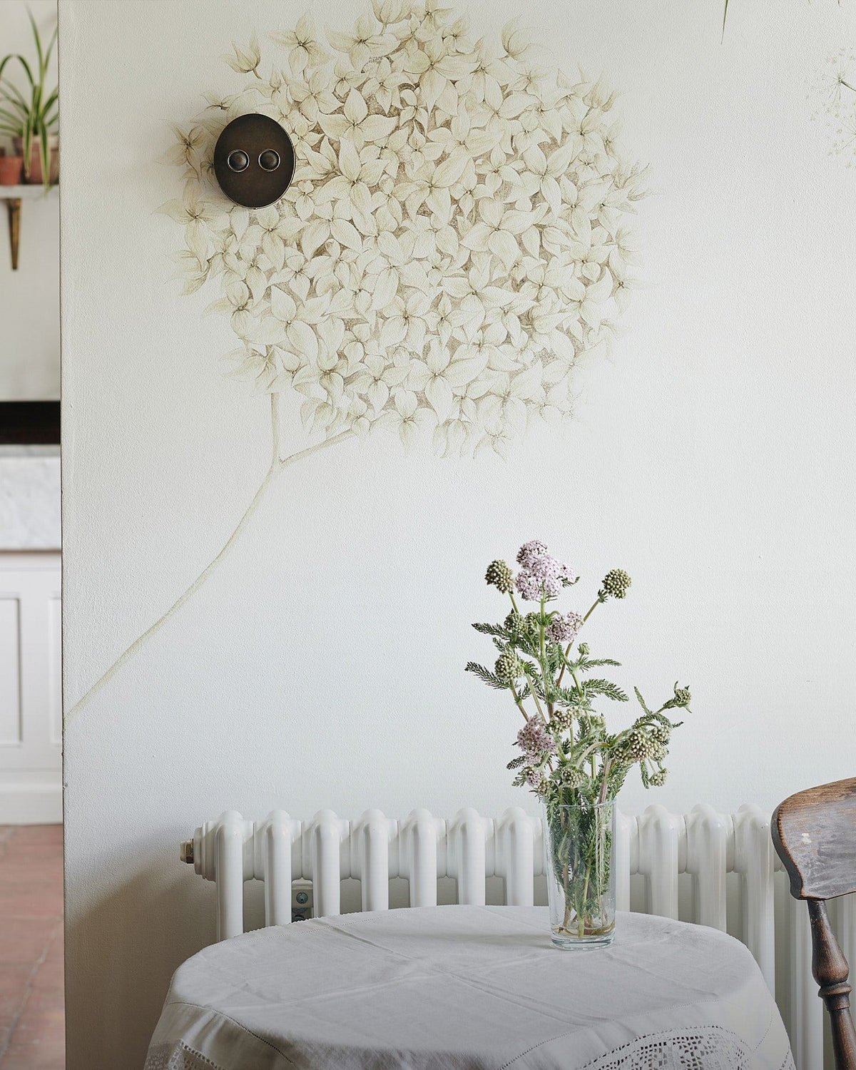 A straight-on view of a 2 gang oval-shaped dimmer switch in a dark oxidised brass finish on a white wall with a large mural of an allium flower head. A small round table with a white linen tablecloth and a vase of flowers is in the foreground.  TEST