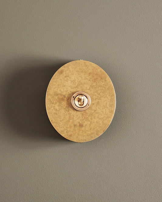 A straight-on view of a 1 gang oval-shaped toggle switch in a mottled aged brass finish with a black base.
