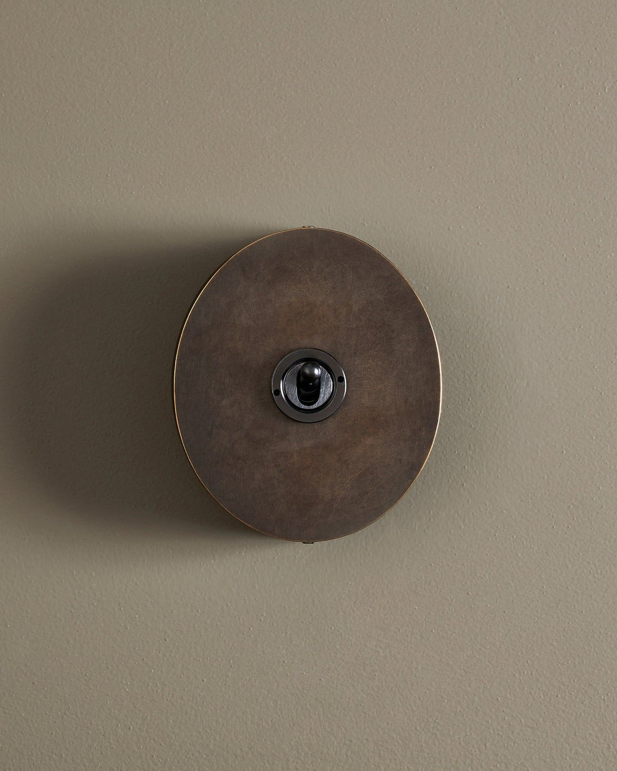 A straight-on view of a 1 gang oval-shaped toggle switch in a dark oxidised brass finish with a black base. TEST